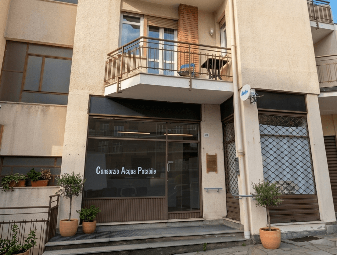Ingresso dell'ufficio Consorzio Acqua Potabile con vetrina, piante in vaso e un balcone sovrastante.